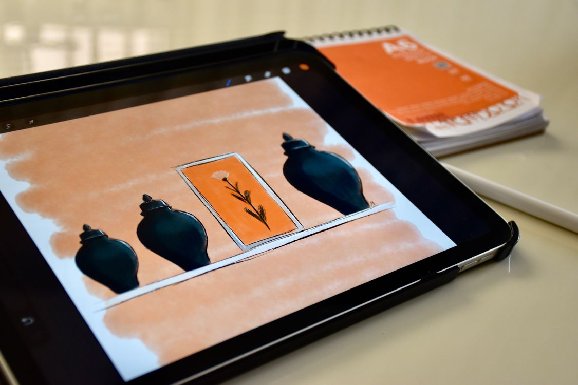Close photograph of a tablet displaying a vase-and-flower sketch beside a stylus and paper notebook.