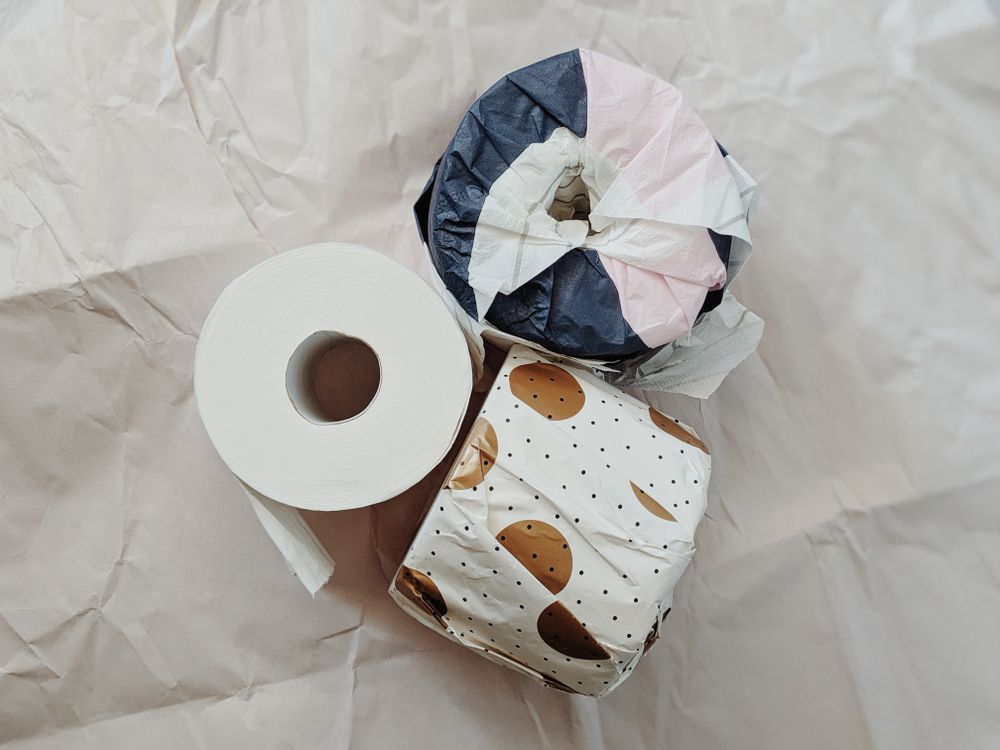 Still-life photograph of toilet-paper rolls wrapped in printed abstract geometric packaging on a neutral surface.