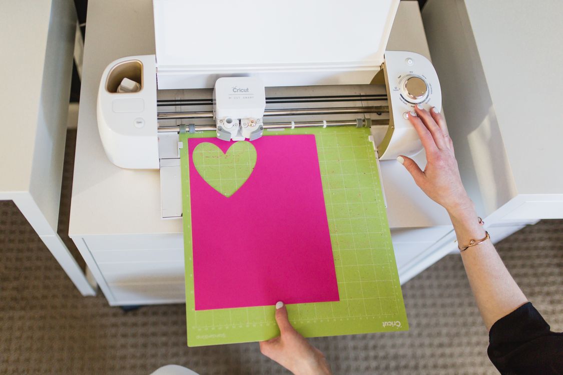 Overhead photograph of a person feeding pink paper into a desktop cutting machine as it cuts a heart shape.