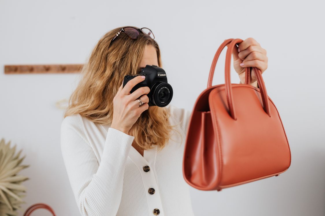 Woman photographing a brown handbag with a camera in a small indoor product-shoot setup.