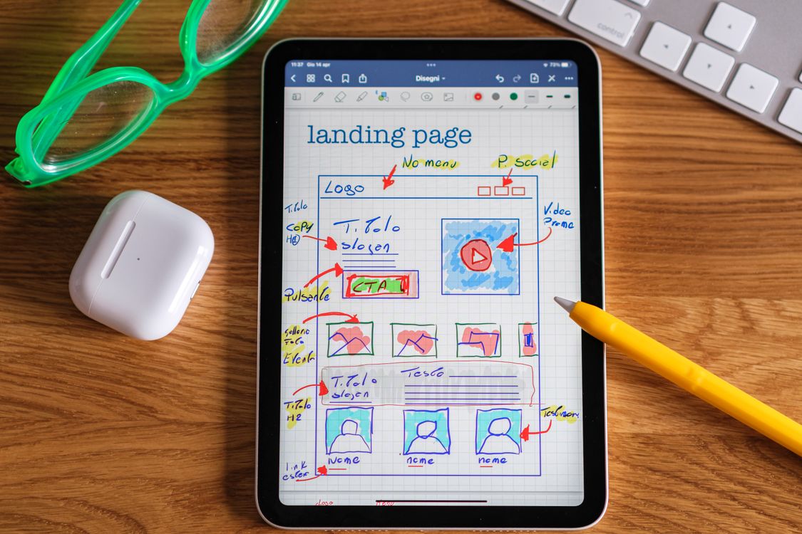 Close-up of a tablet showing a hand-drawn landing-page layout beside a keyboard, pencil and green glasses.
