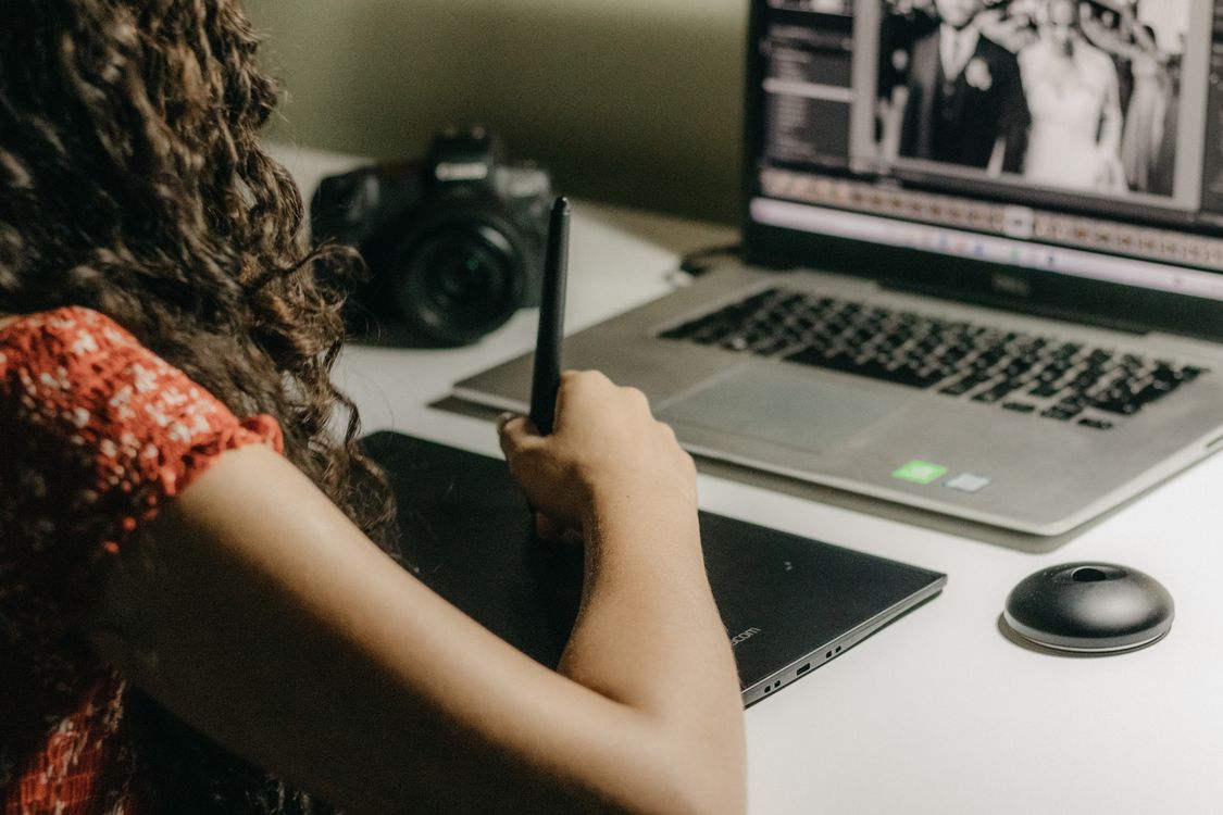 Rear view of a woman editing photographs on a laptop beside a pen tablet at a desk.